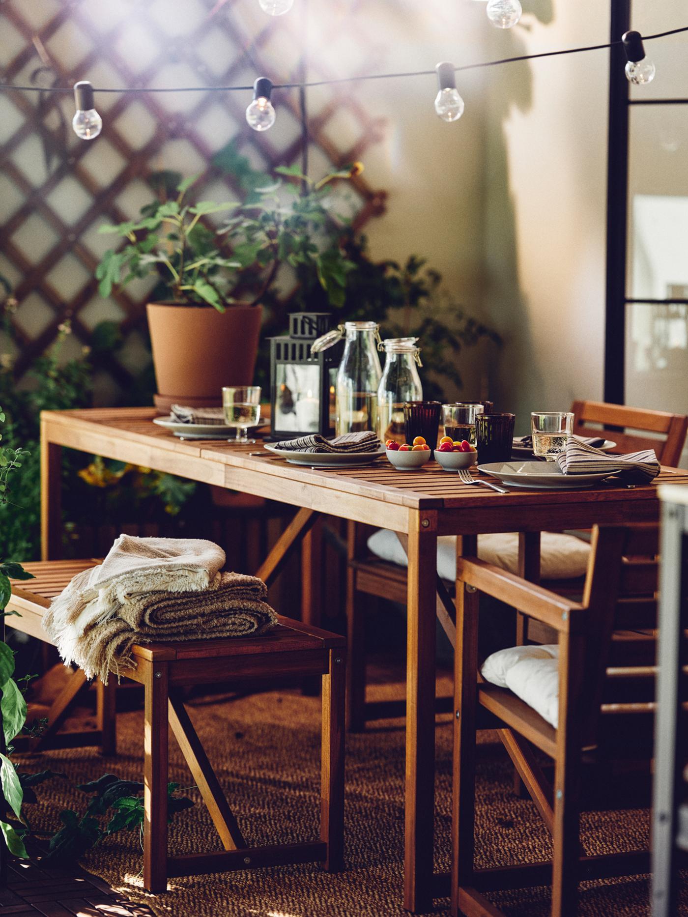 A patio with a NÄMMARÖ outdoor table, bench and chairs with armrests. A SVARTRÅ LED lighting chain hangs above the table.