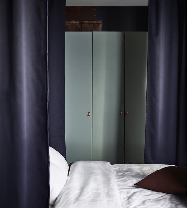 A PAX wardrobe with hinged grey-green doors in a bedroom with dark curtains around the bed.