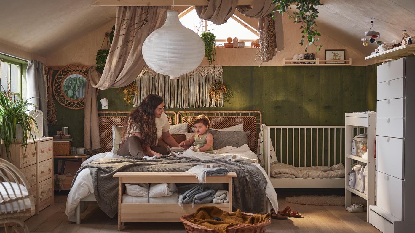 A person and child interacting on a VEVELSTAD bed that’s covered in neutral-coloured textiles beside a white GULLIVER cot.