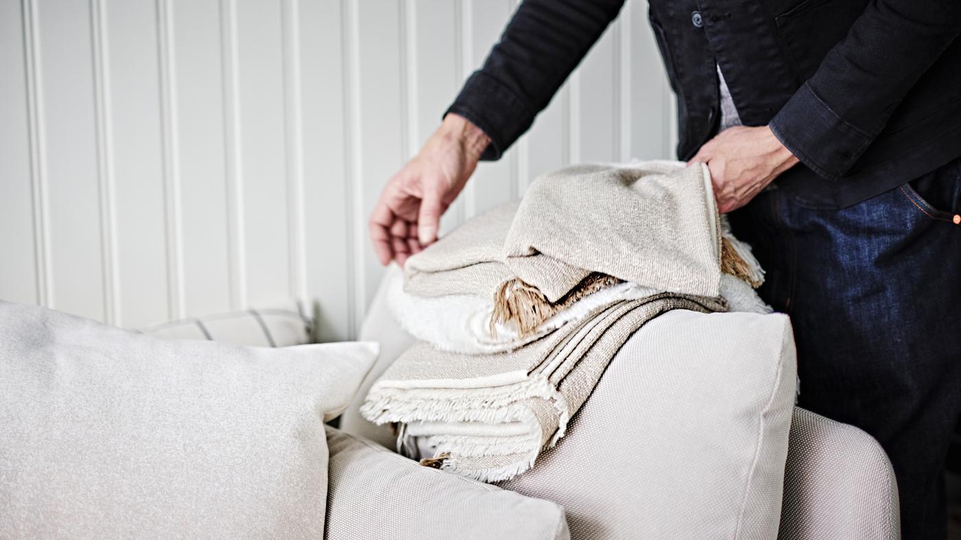 A person folding a grey-beige GUNNEMOR throw on the arm of a light beige SÖDERHAMN 1-seat corner section.
