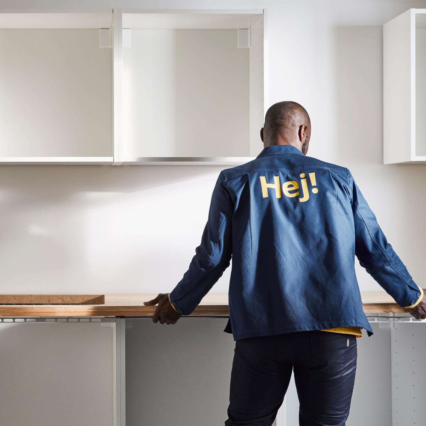A person in a blue jacket installing a white kitchen with base and top cabinets and an oak effect/laminate worktop.