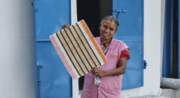 A person in a pink vest stands before a weathered white wall and blue window shutters. They are smiling and holding a striped MÄVINN place mat in their hands.