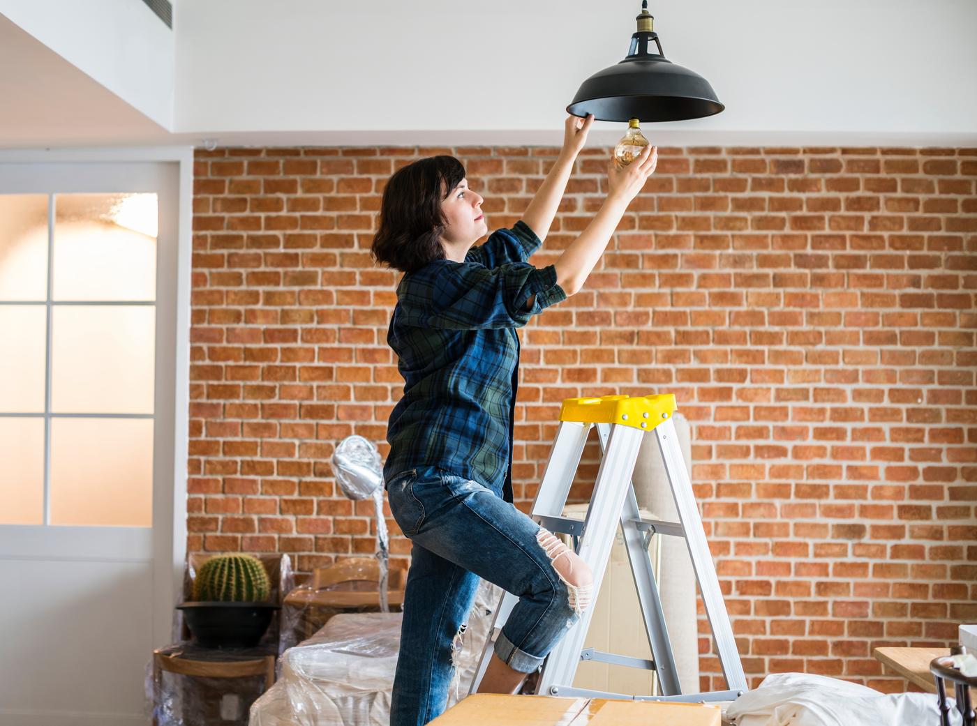 A person is changing a light bulb on a pendant lamp shade, whilst standing on a ladder inside a house that is being renovated.