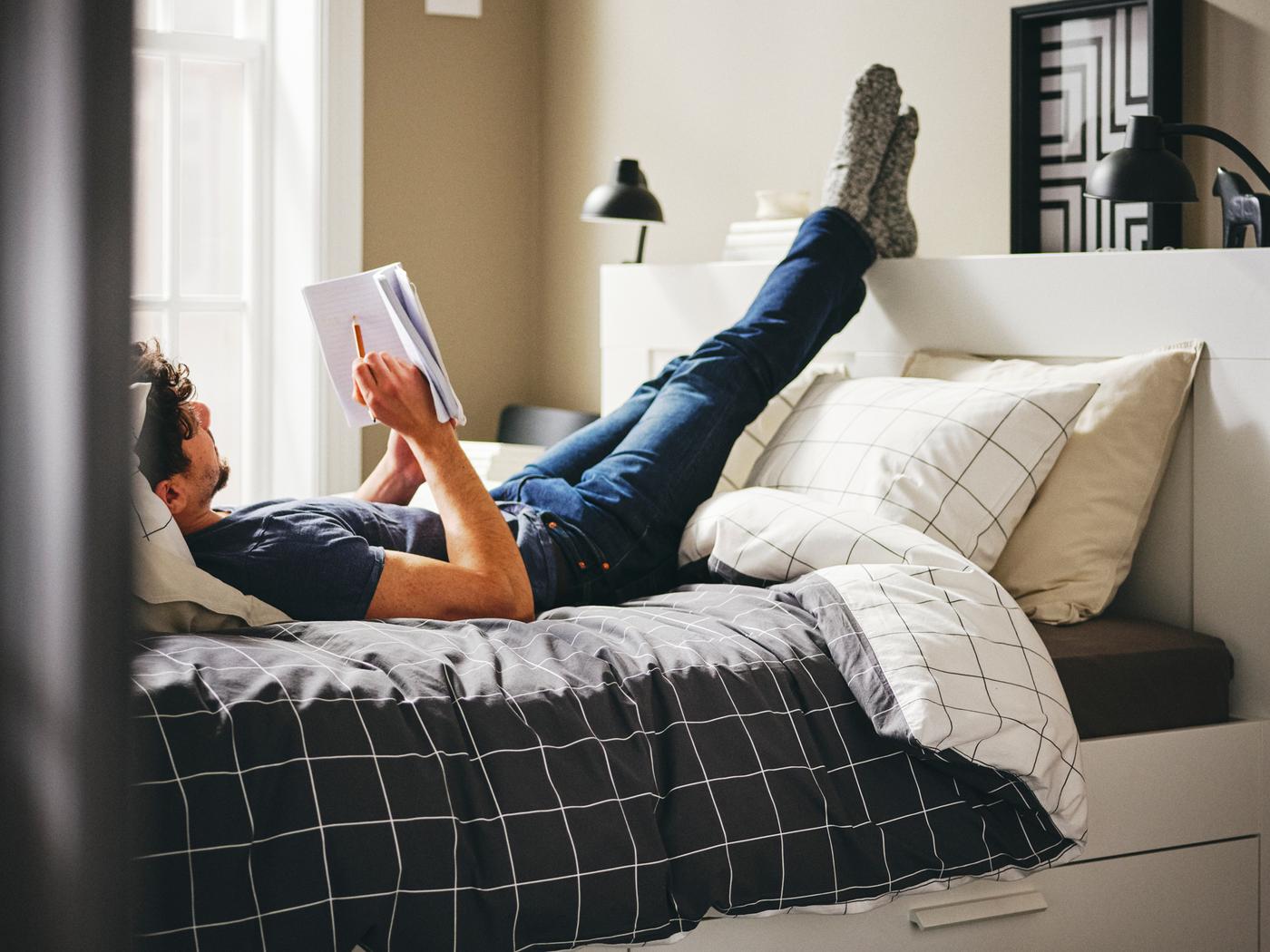 A person lies on a white BRIMNES bed with storage and headboard, with their feet on the headboard, holding a book and pencil.