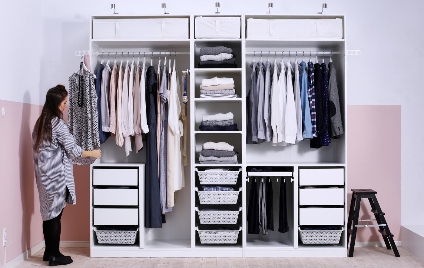 A person places a shirt to hang at the side of a large white wardrobe with rails, drawers, shelves and no doors.