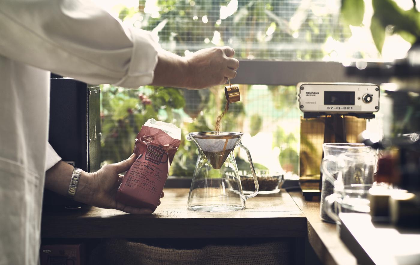 A person pours ground coffee into a filter on a wooden worktop with greenery outside the window behind.