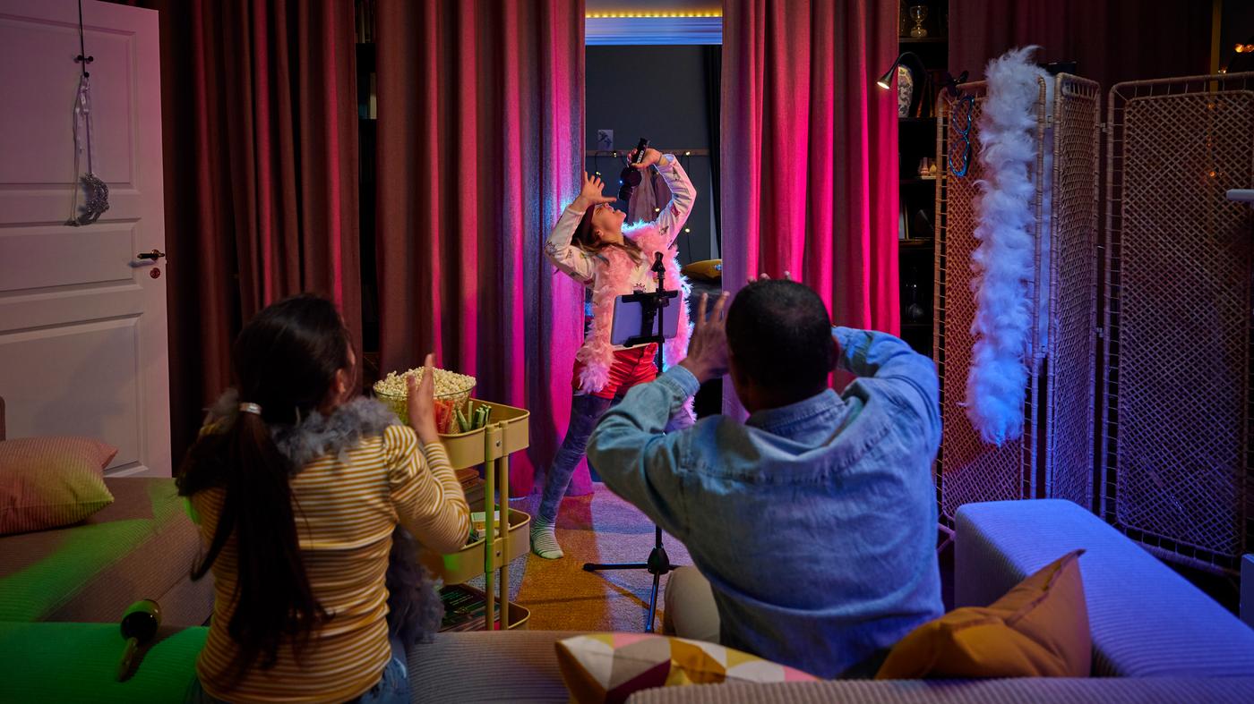A person singing in front of a dark red curtain, with two persons in a grey/beige sofa with chaise longues looking on.