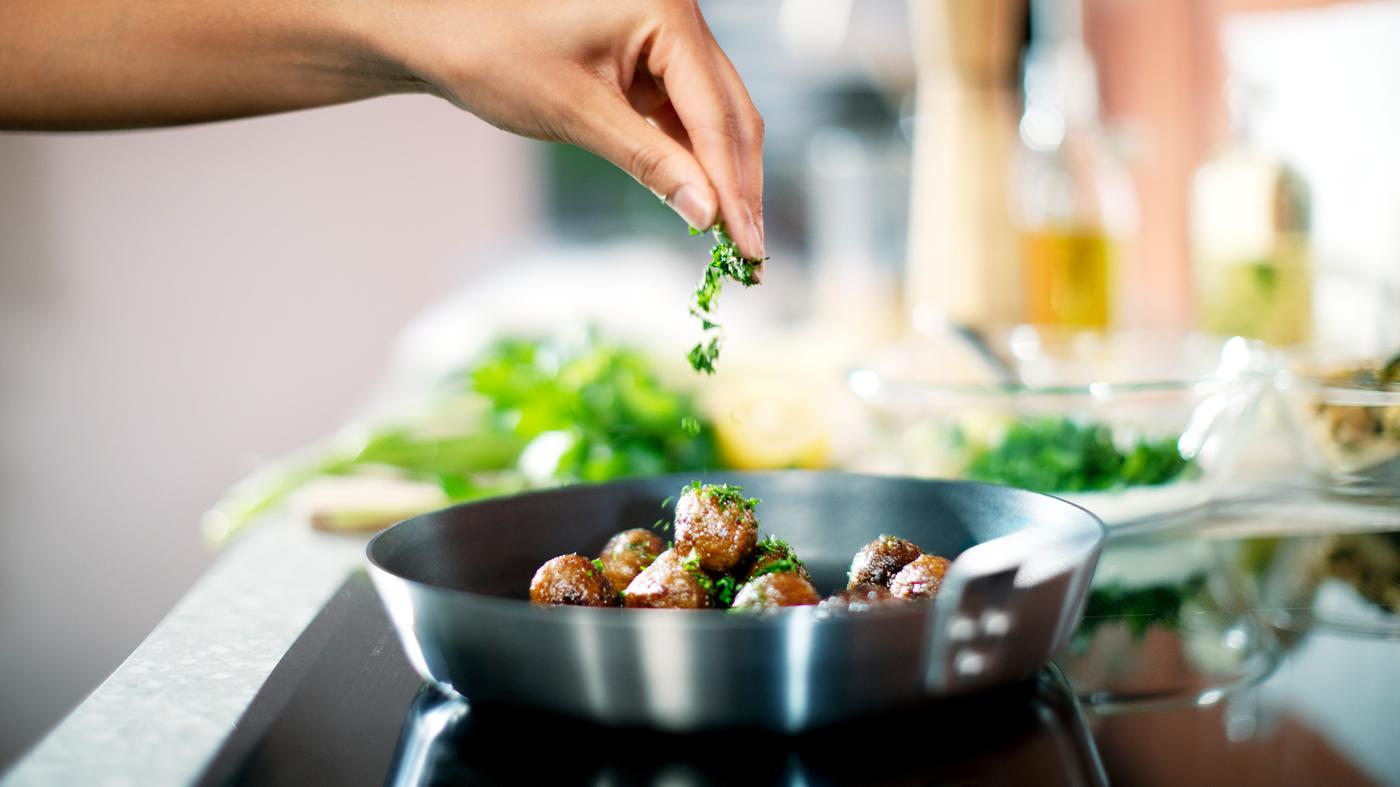 A person sprinkles chopped green herbs on to HUVUDROLL plant balls in a IKEA 365+ frying pan on a hob.