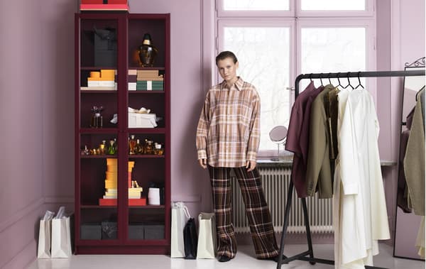 A person standing between a glass-fronted book case and an open rail of clothes.