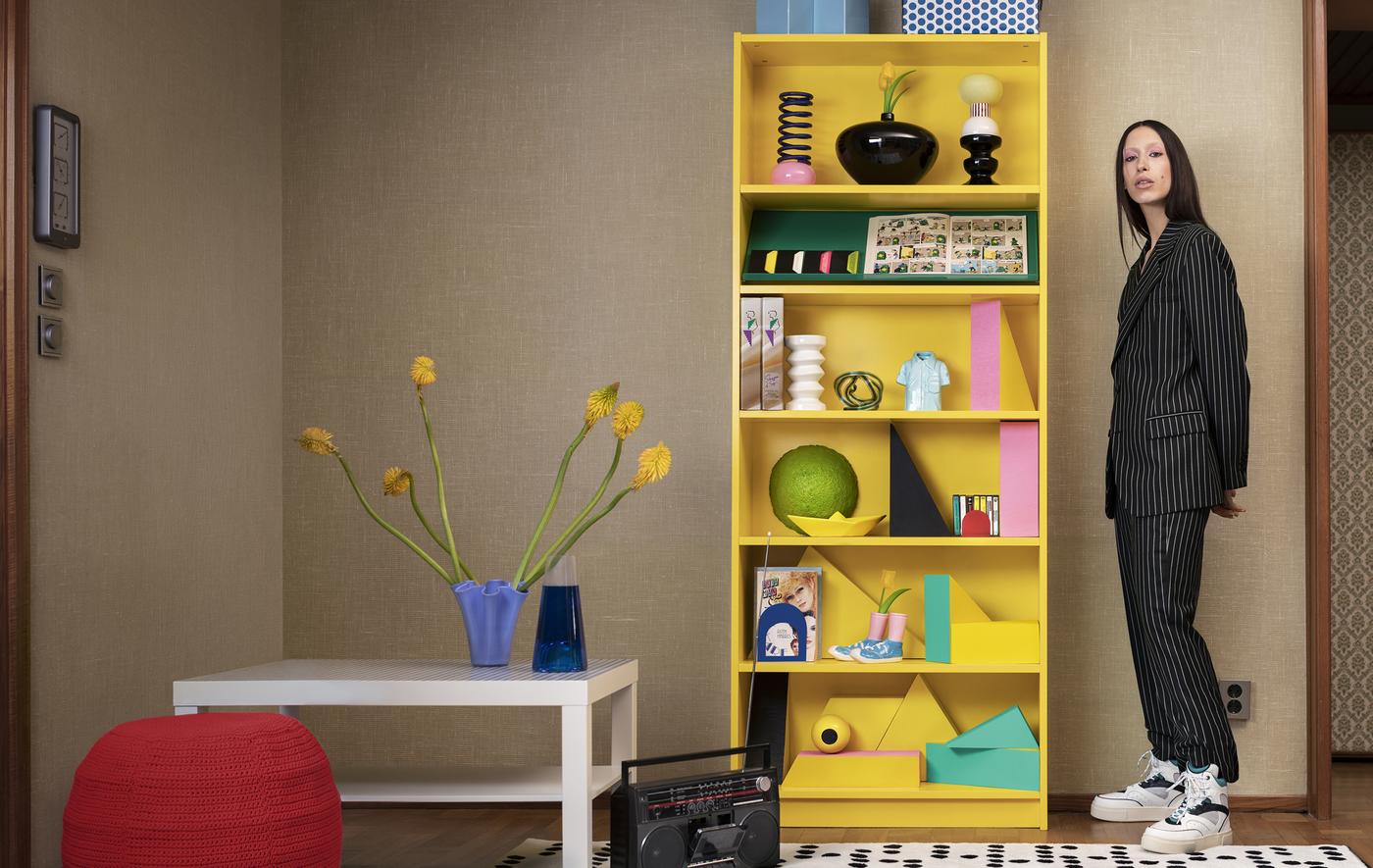 A person standing next to a bright yellow BILLY bookcase with ornaments on the shelves.