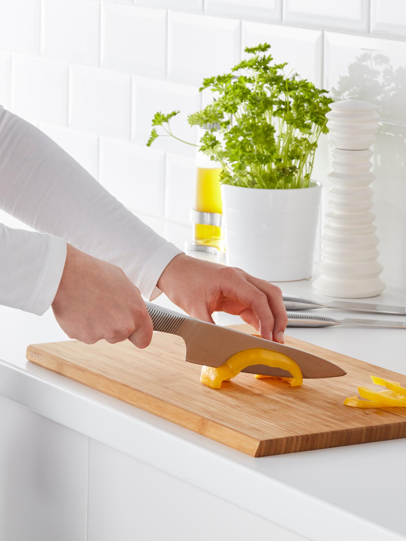 A person wearing a white long sleeve shirt is slicing a yellow capsicum on a chopping board using an IKEA 365+ cook’s knife.