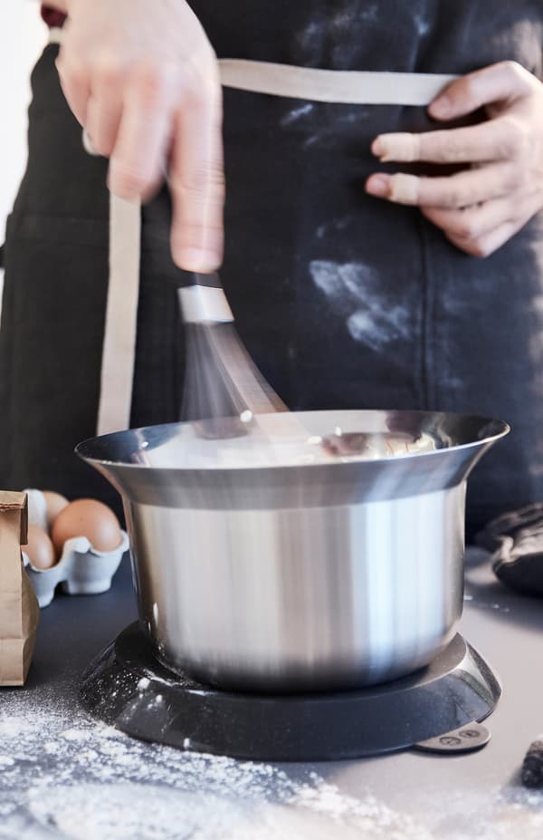 A person whisks with one hand. OMTÄNKSAM mixing bowl has a lid that can be placed underneath to stop it from slipping.