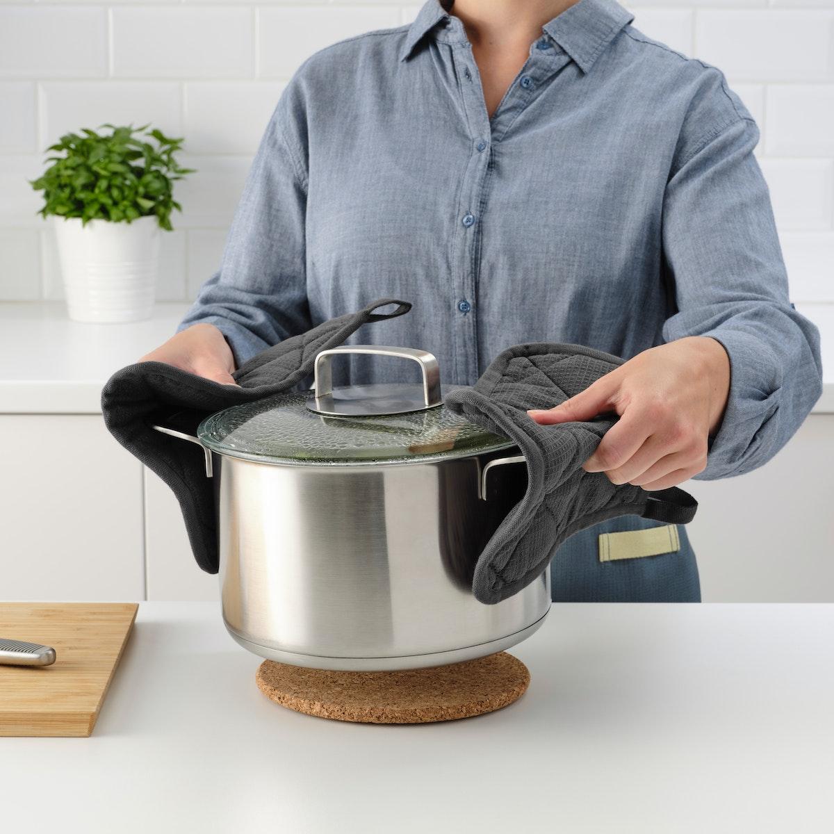 A person with a light blue shirt uses RINNIG pot holders hold sides of large pot and place it onto a cork trivet. 