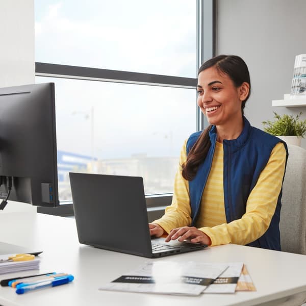 A person with IKEA co-worker shirt sits at white desk working on computer behind window.