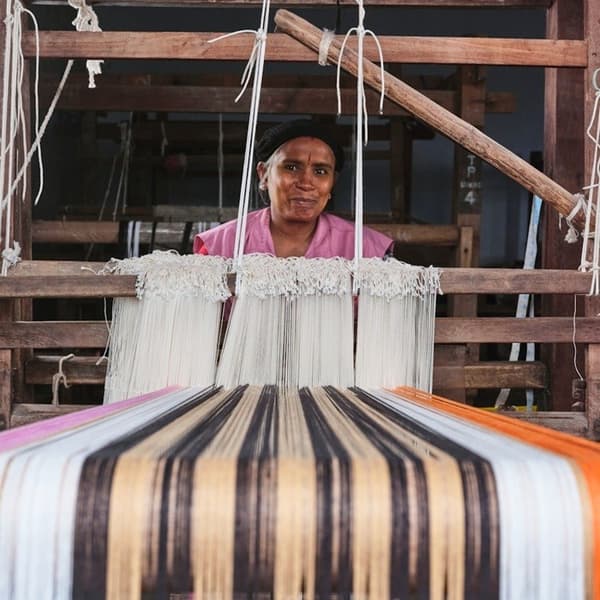 A person working at a handloom, weaving a striped MÄVINN table-runner in shades of beige, black, pink and orange.