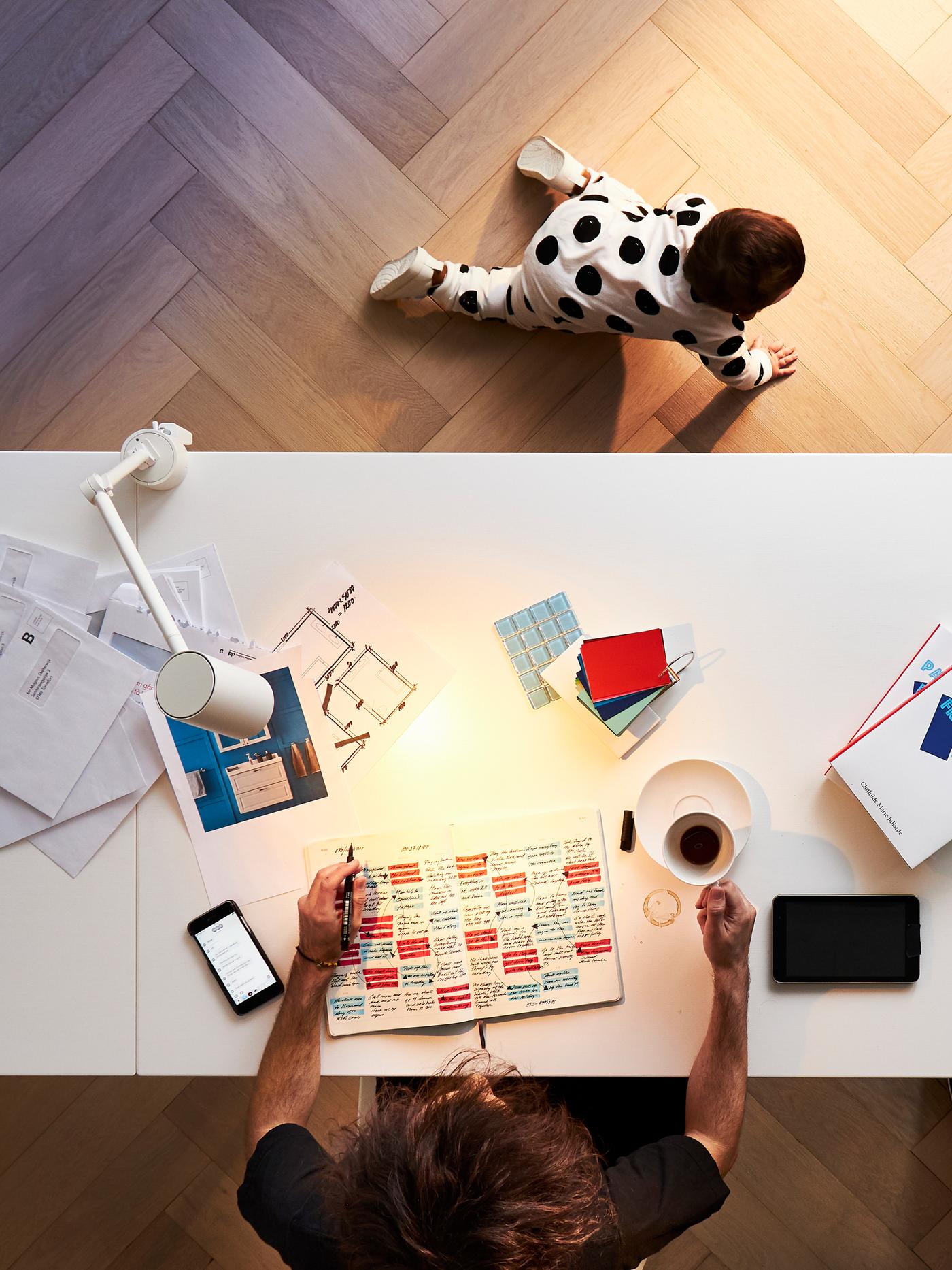 A person works at a home office desk with a cup of coffee and a pen and paper while a baby crawls on the floor nearby.
