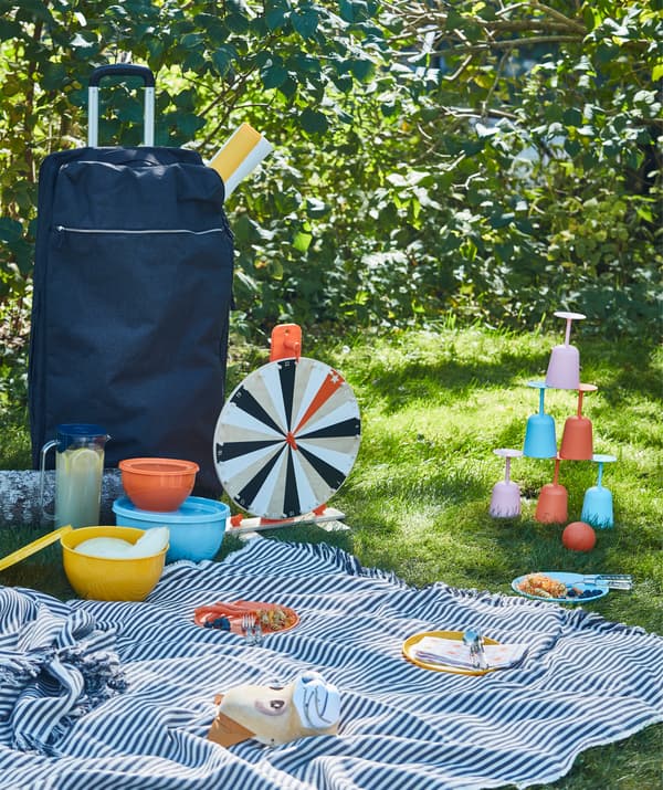 A picnic in the grass with tableware, games and a big blanket brought with a blue FÖRENKLA XL duffle bag on wheels.