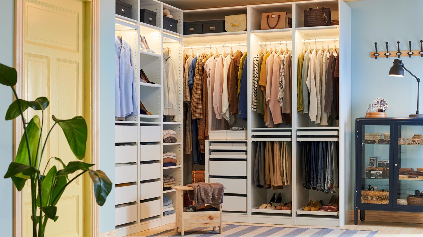 A pine PERJOHAN stool with storage stands in front of a large open PAX corner wardrobe with clothes, shoes, boxes and bags.