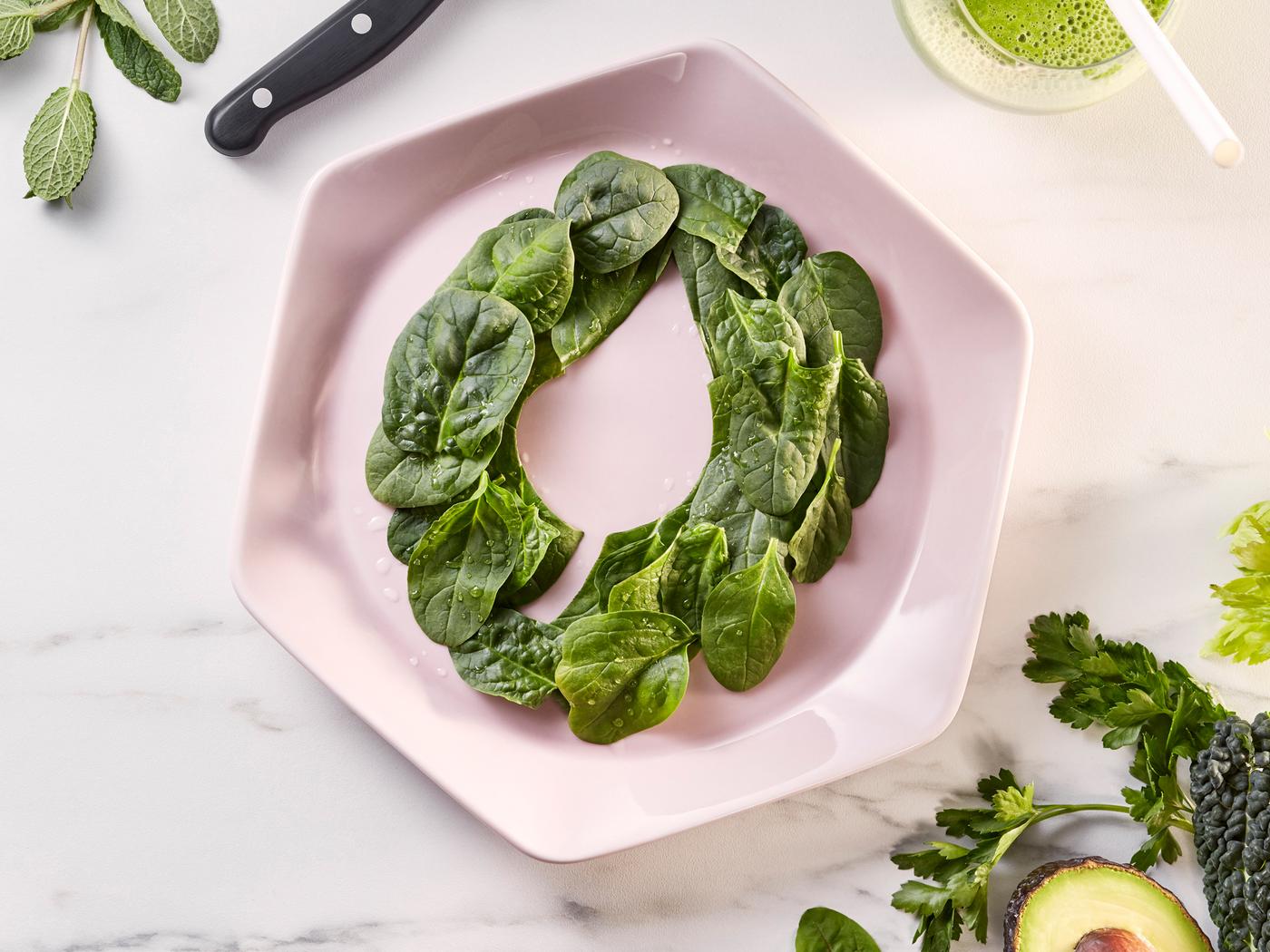 A pink hexagonal plate on white marble bench top. On the plate are baby spinach leaves decoratively arranged in a ring.