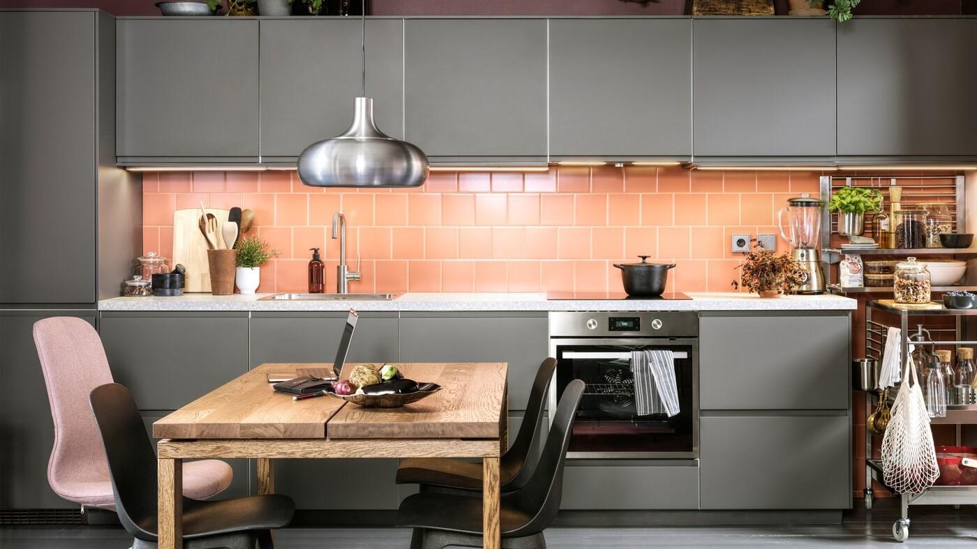 A pink-tiled kitchen with dark grey kitchen fronts, anthracite chairs, a dining table in oak veneer and a VÄXJÖ pendant lamp.