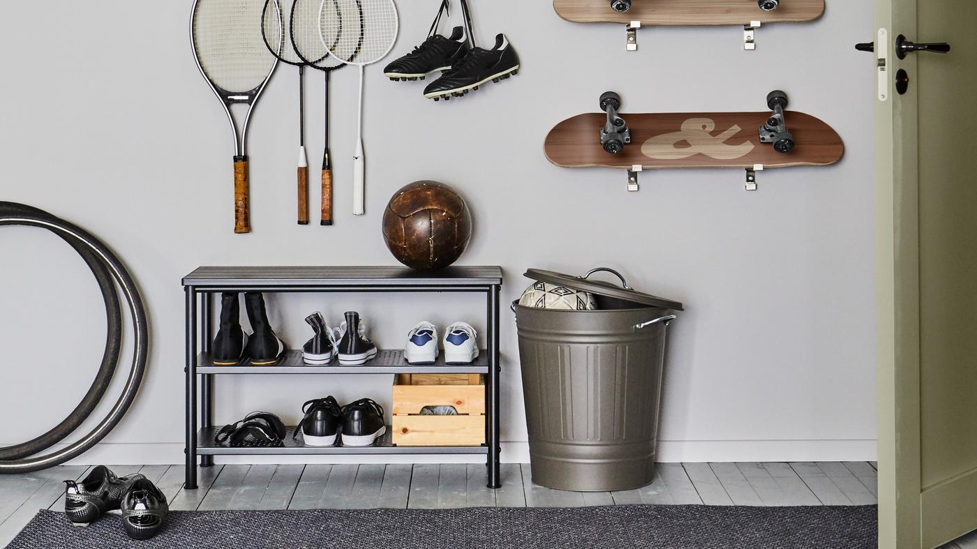 A PINNIG shoe rack with shoes and a football by a KNODD bin with an open lid. Rackets and skateboards are on the wall above.