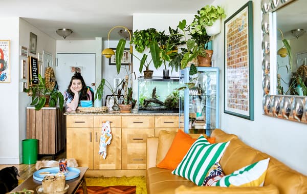 A plant-filled, open-plan living space with a kitchen island, fish tank, sofa, cushions and coffee table, and wall decor.