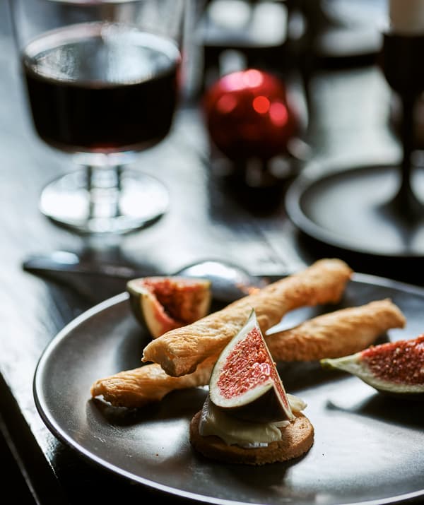 A plate of party food with fig, cheese, biscuits and breadsticks, on a table with a glass of wine and red Christmas bauble.