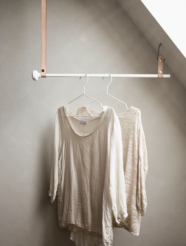 A pole hung from leather straps attached to a sloped ceiling holding clothing on hangers.