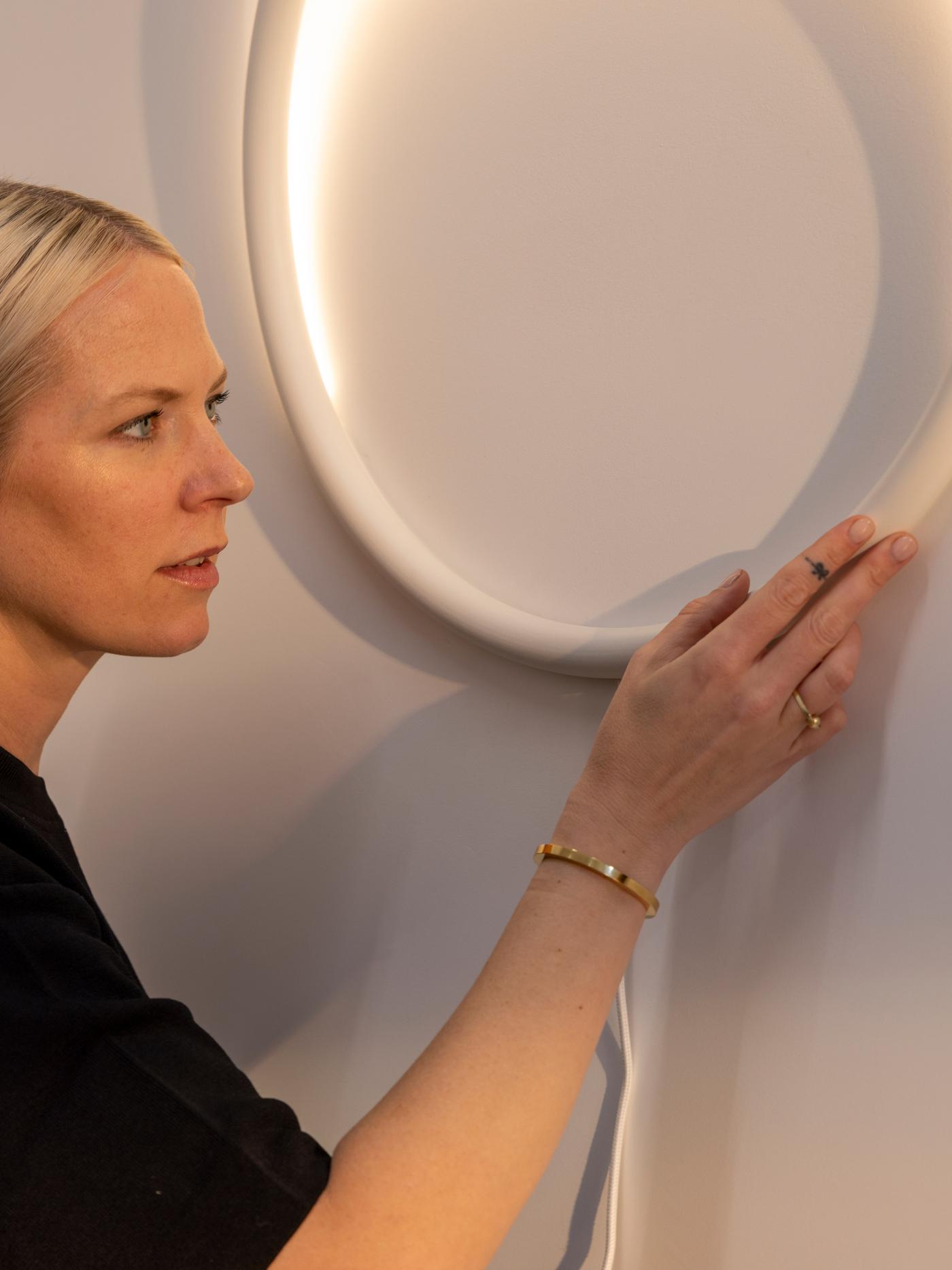 A portrait of designer Sabine Marcelis, dressed in black, inspecting a prototype of a VARMBLIXT circular wall lamp.