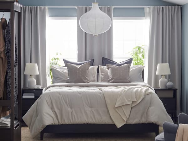 A primary bedroom with gray curtains and a white lampshade above a neatly made bed with decorative pillows on it.