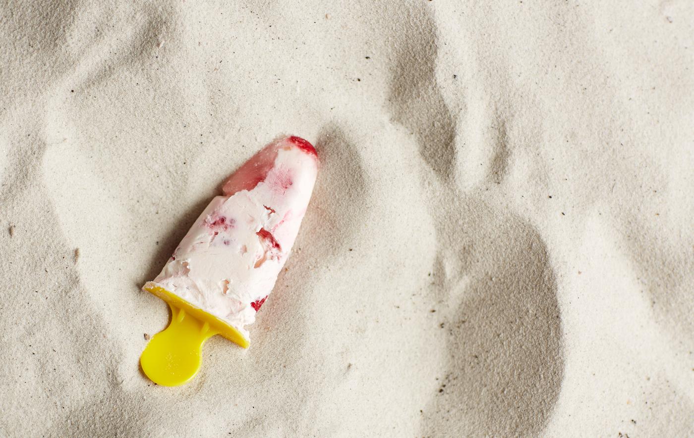 A raspberry frozen yogurt pop lies in the sand.