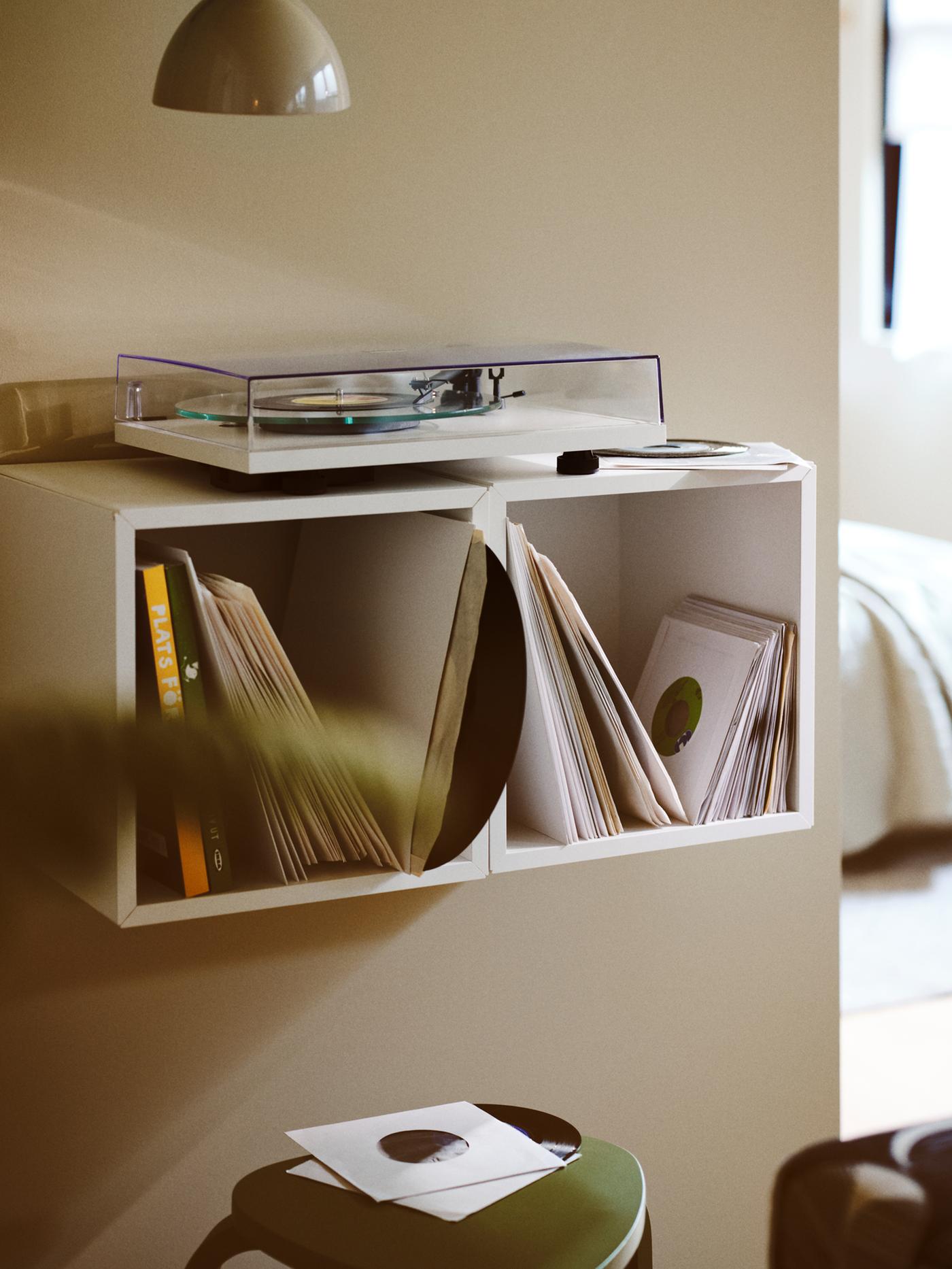 A record player placed on top of two side-by-side, white EKET wall-mounted shelving units holding multiple vinyl records.