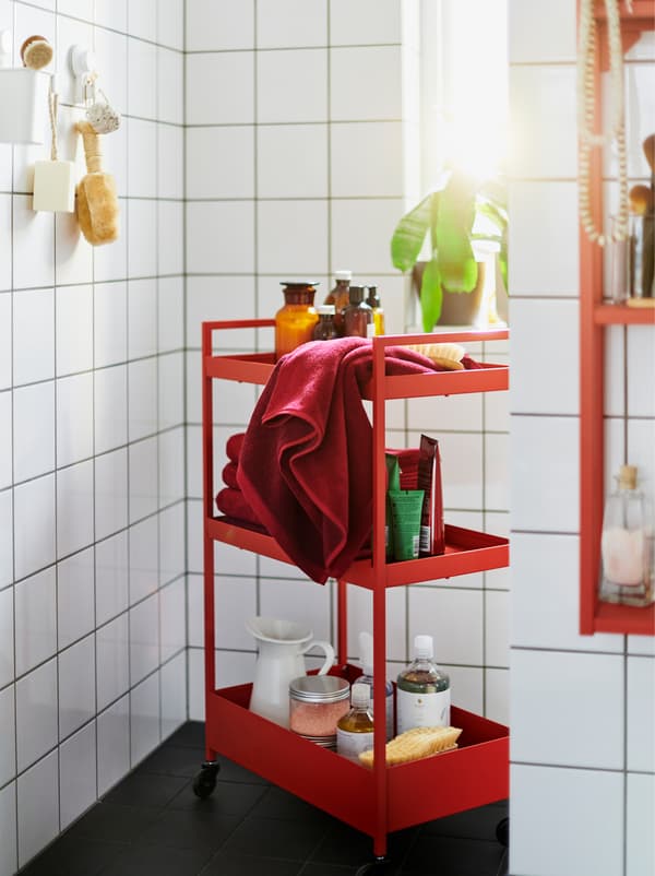 A red NISSAFORS trolley filled with accessories in a white-tiled bathroom also furnished with red and white ENHET modules.