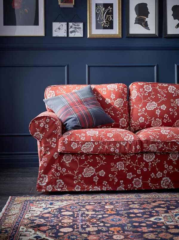 A red, patterned EKTORP sofa with a plaid cushion in a living room with dark, panelled walls covered with framed art.