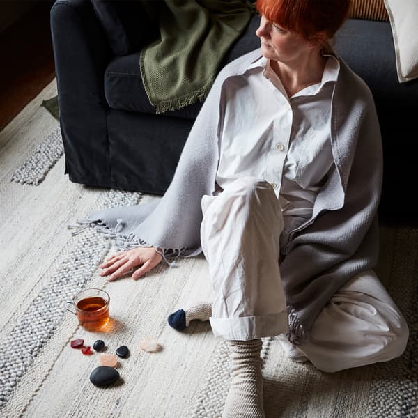 A redheaded woman sitting on a rug with a shawl over her shoulders, leaning on the sofa, beside a glass of tea and decorative stones.