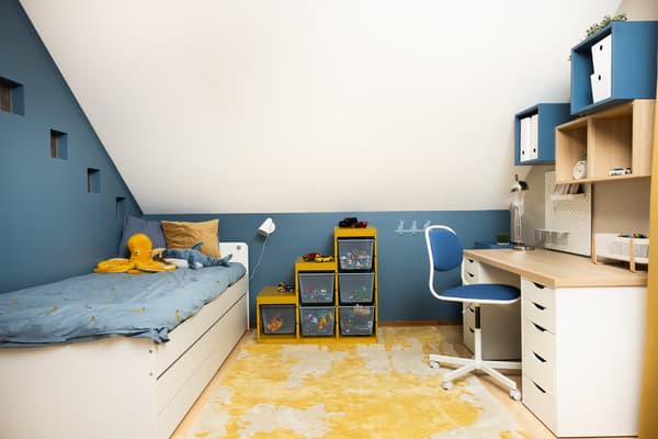 A renovated children’s room with a bed under a sloped ceiling, a study desk with chair, wall-mounted shelving, and colorful storage boxes. Blue and yellow accents, light wood surfaces, and defined zones.