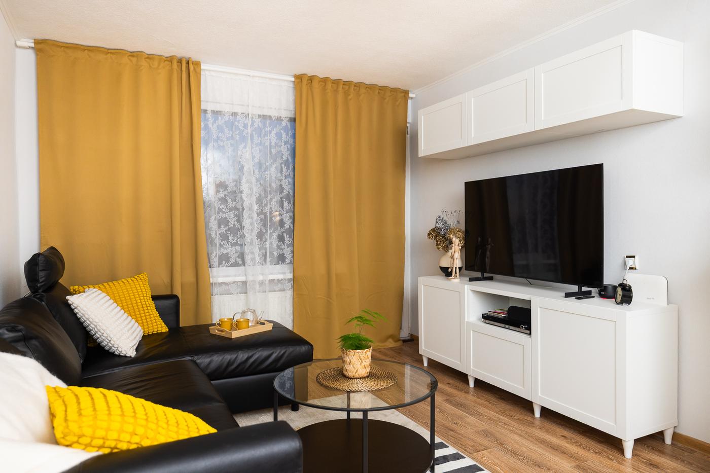 A renovated living room with a black sofa, a white TV unit, and mustard-yellow curtains. The space looks tidy, bright, and functionally arranged.