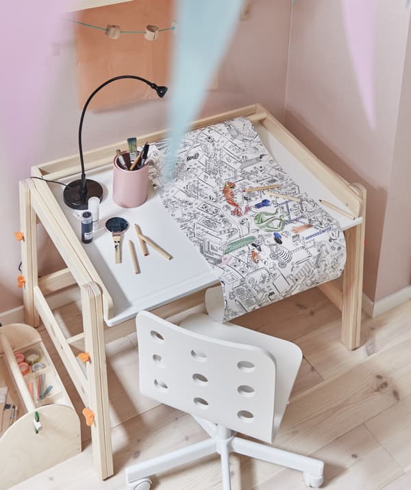 A roll of colouring paper on a child’s desk with a white chair against a pink wall.