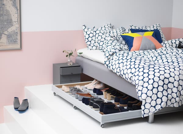 A rolling box that fits under the bed in this white and pink bedroom is filled with shoes.