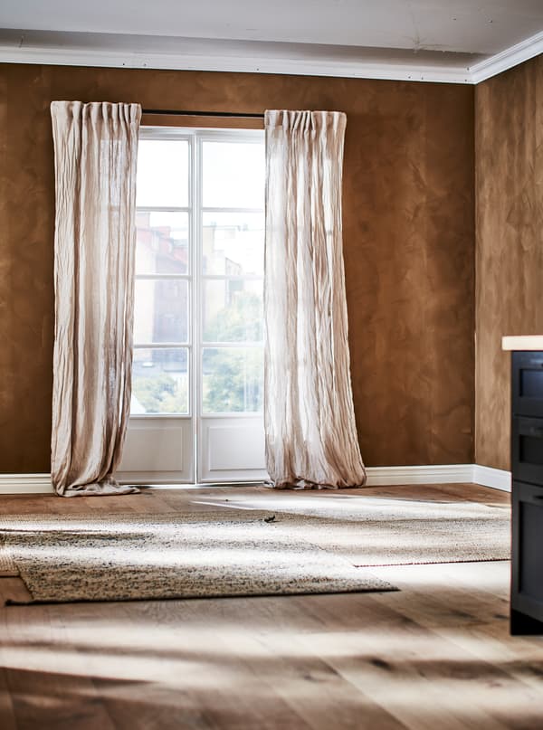 A room painted brown, with French windows dressed with crinkled-linen curtains, and light-brown, natural-material rugs on the wooden floor.