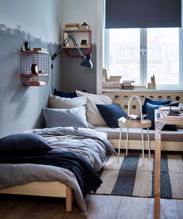 A room with a corner sofa and single bed, with textiles in dark blue, grey and natural colours and red wire wall storage.