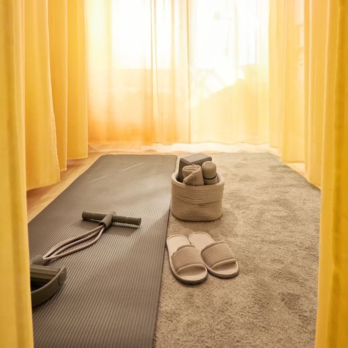 A room with a yoga mat and slippers on the floor, yellow curtains all around.