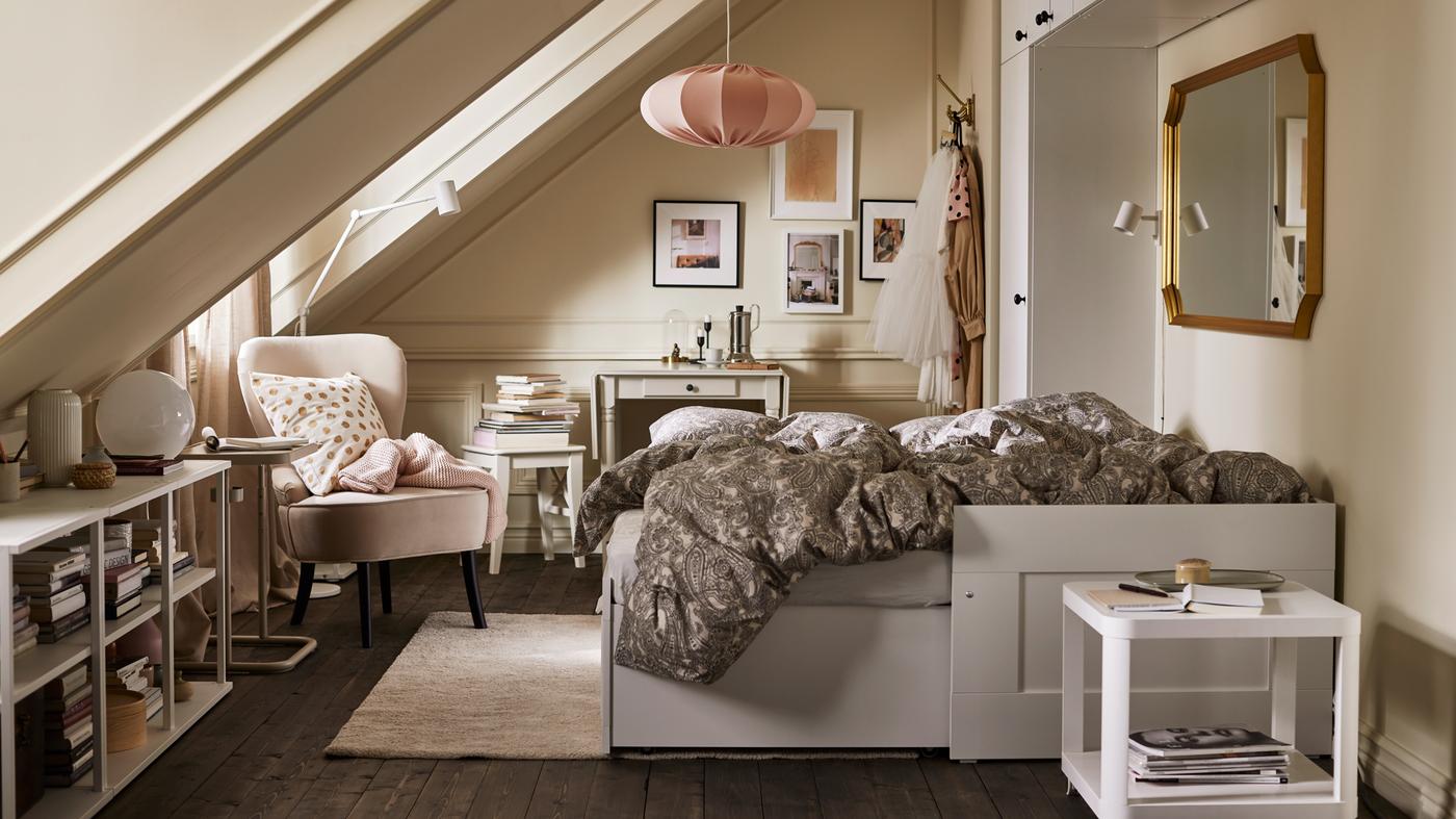 A room with an extended white daybed with beige/dark grey bed linen, various storage, a beige armchair and pink pendant.