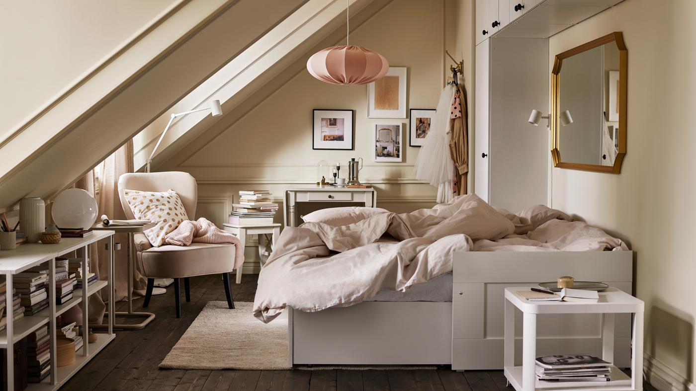 A room with an extended white daybed with light gray/beige bed linen, various storage, a beige armchair and pink pendant.
