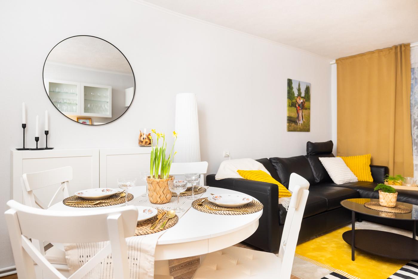 A round dining table with white chairs beside a wall with a large round mirror. The space appears bright, tidy, and visually divided into functional zones.