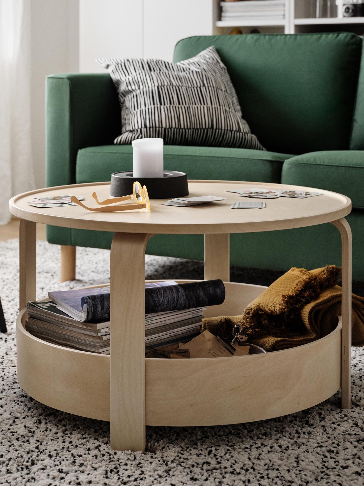A round light wood BORGEBY table in front of a green sofa with playing cards, a block candle and a pair of spectacles on top.