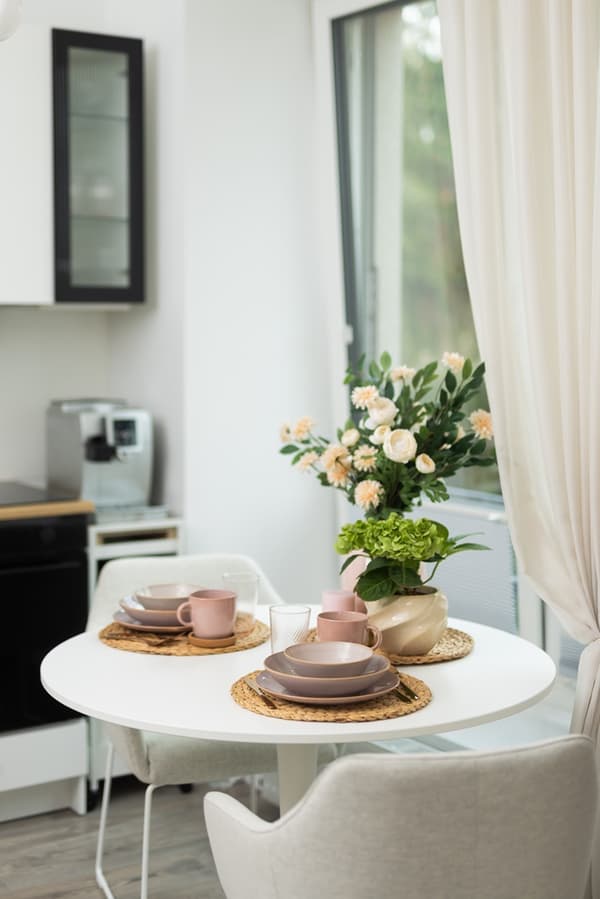 A round table by the window set with tableware, flowers in a vase, and curtains creating a cosy atmosphere.