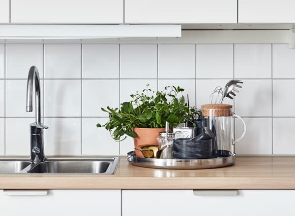 A round tray holding potted herbs, kitchen utensils and a carafe sits on a kitchen counter.