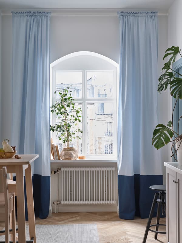 A rounded kitchen window with a large plant on the windowsill and light blue BENGTA block-out curtains on both sides.