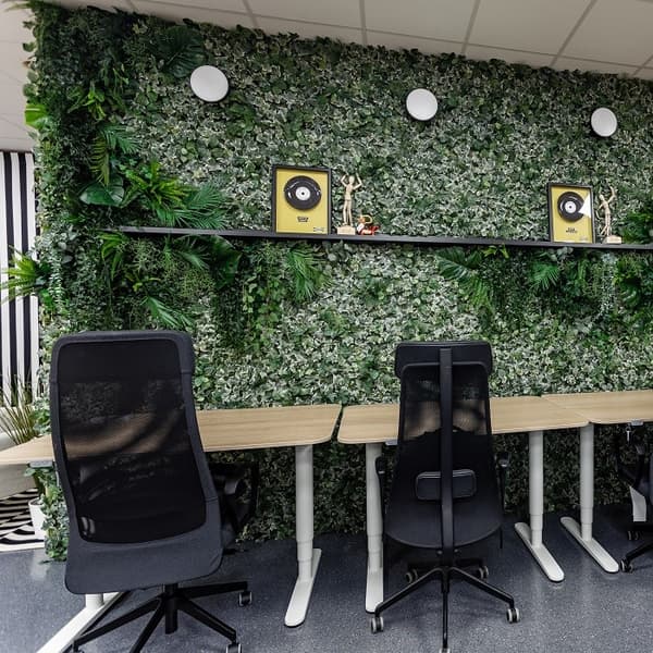 A row of modern office desks and ergonomic black chairs faces a vibrant green wall covered entirely with artificial ivy and foliage, creating a lush, biophilic design. A long black floating shelf spans the wall, displaying framed gold records, small decorative figurines, and minimalist sculptures. Above the shelf, three round white wall lights provide illumination. The scene blends nature-inspired decor with a clean, organized workspace aesthetic.